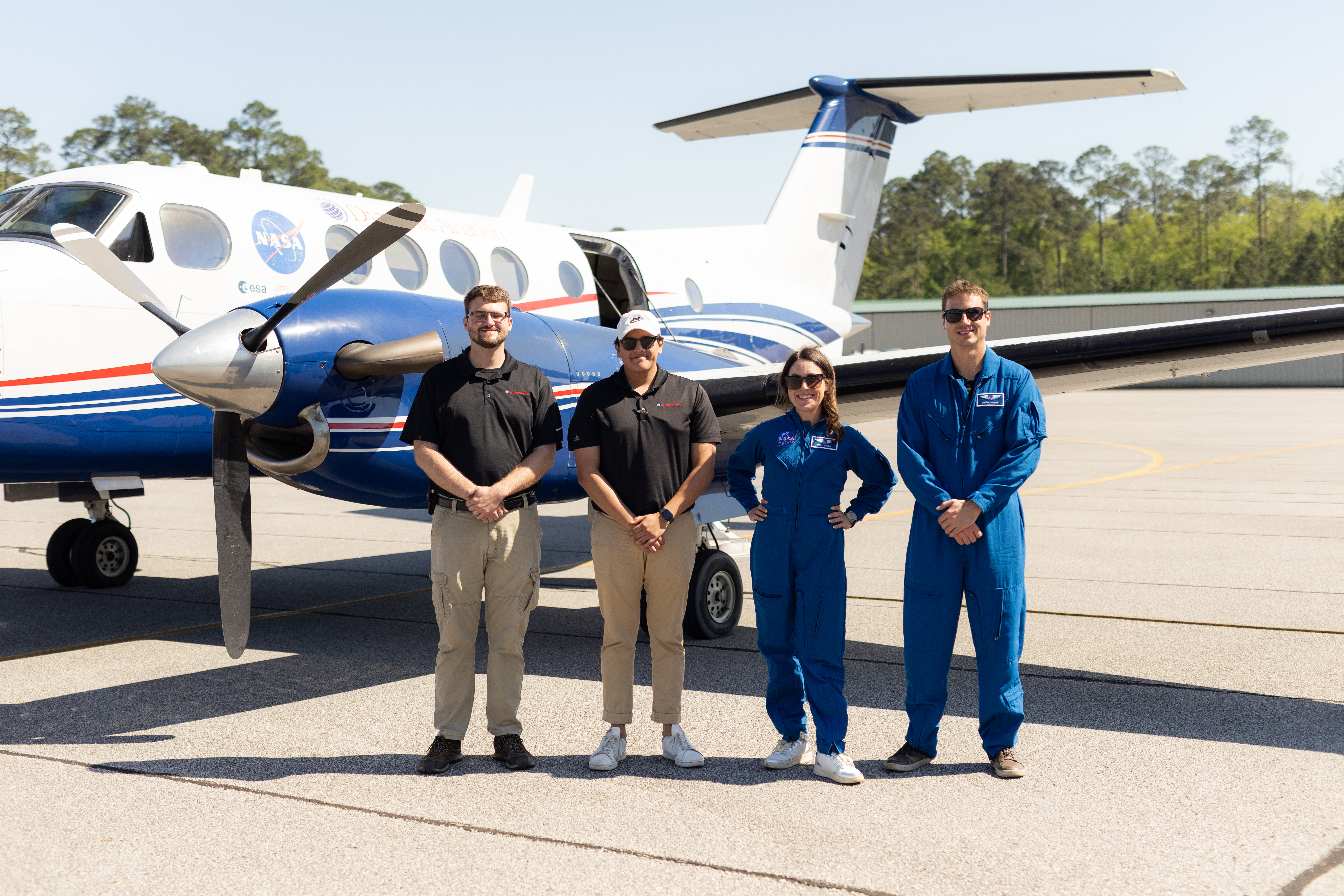 Two pilots in black shirts and tan pants stand beside and two instrument specialists in blue flight suits, in front of a white and blue NASA Armstrong B-200 aircraft on gray tarmac.