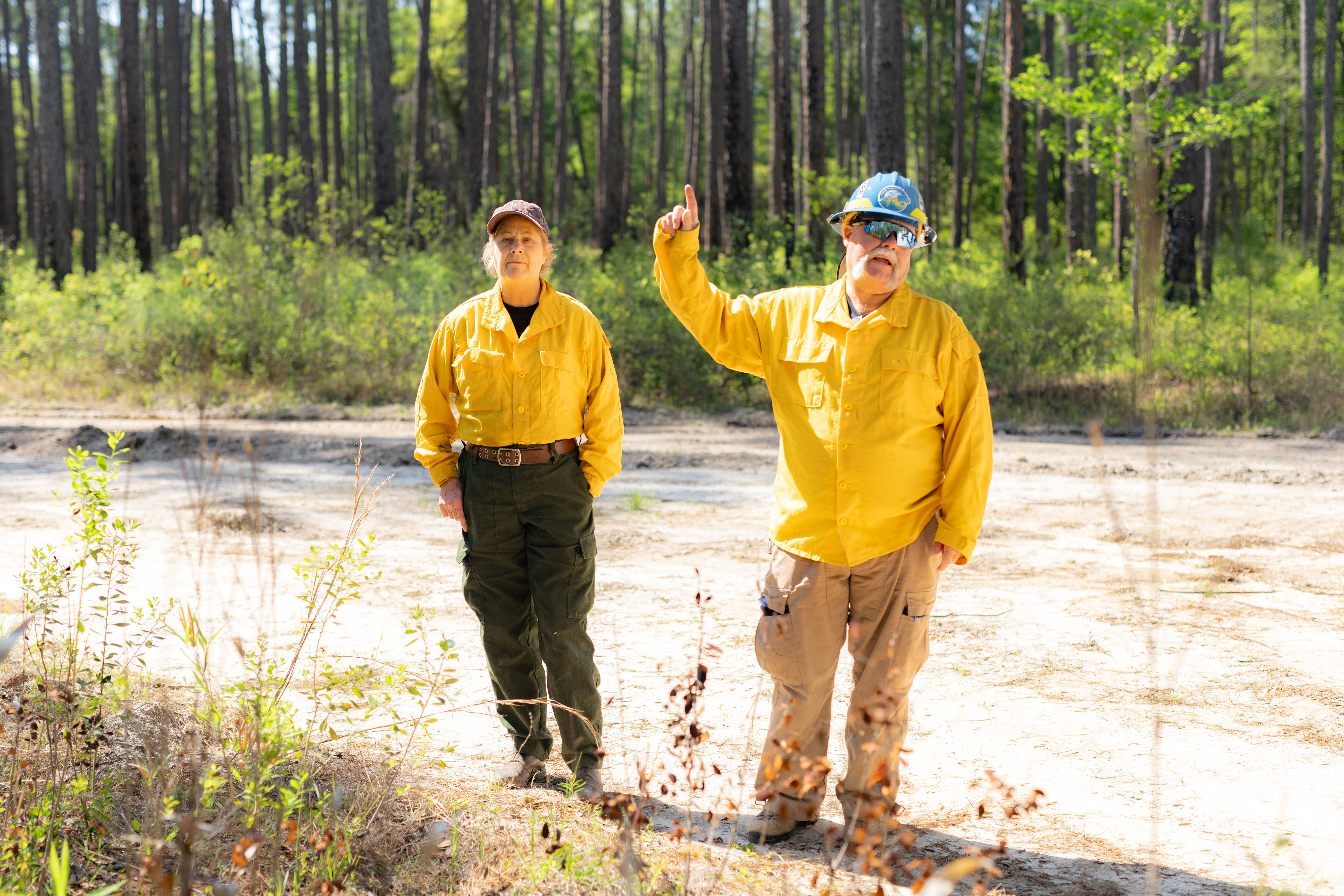 Two safety officers in yellow long-sleeved shirts stand in front of forest, guesturing and speaking.