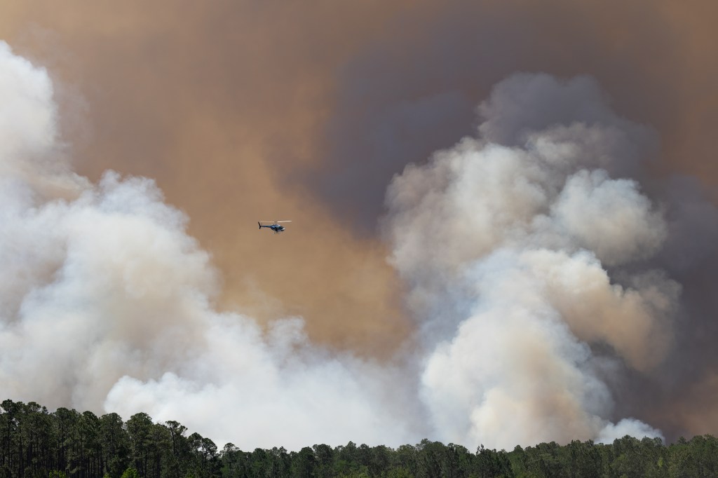 Helicopter flies in front of smokey clouds above a forest.