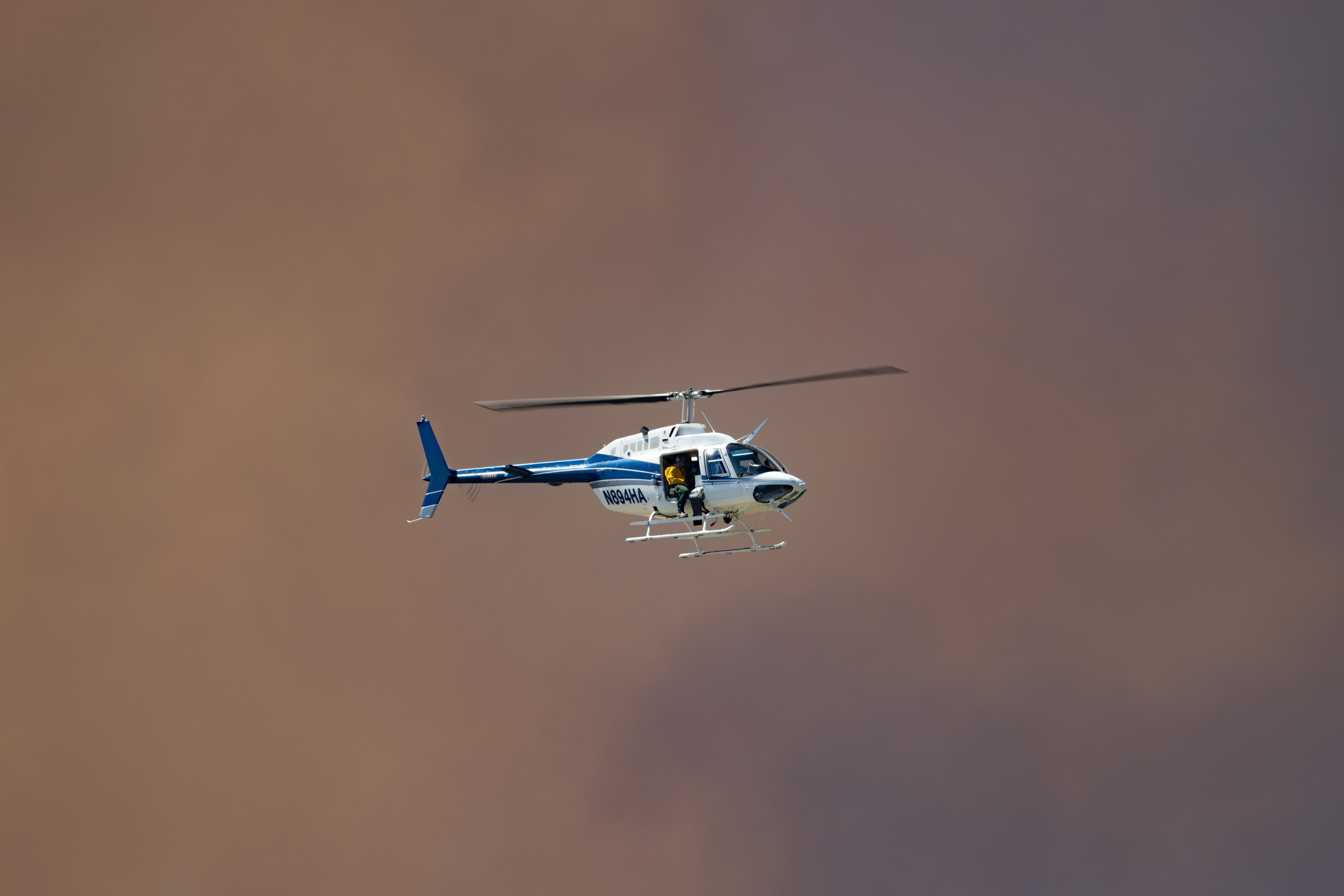 A helicopter, lit by the sun, flies in front of dark pinkish-brown clouds of smoke.