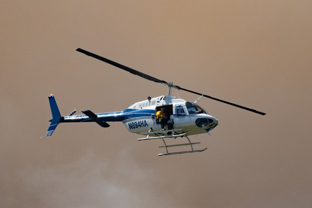 A helicopter, lit by the sun, flies in front of dark pinkish-brown clouds of smoke.