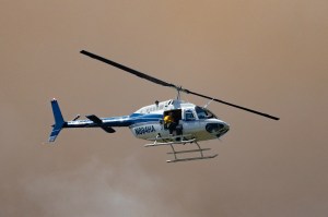 A helicopter, lit by the sun, flies in front of dark pinkish-brown clouds of smoke.
