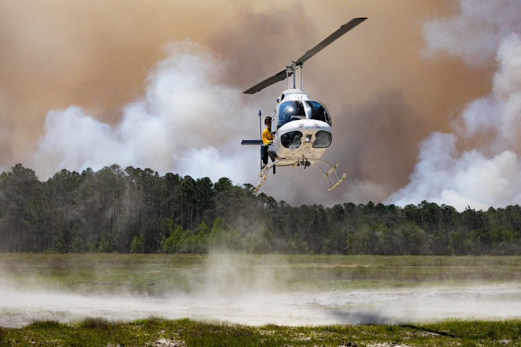 A white helicopter kicks up dust from a grassy clearing as it lands, against a backdrop of dark smoke rising over a green forest. A man in a yellow shirt leans out of the door.