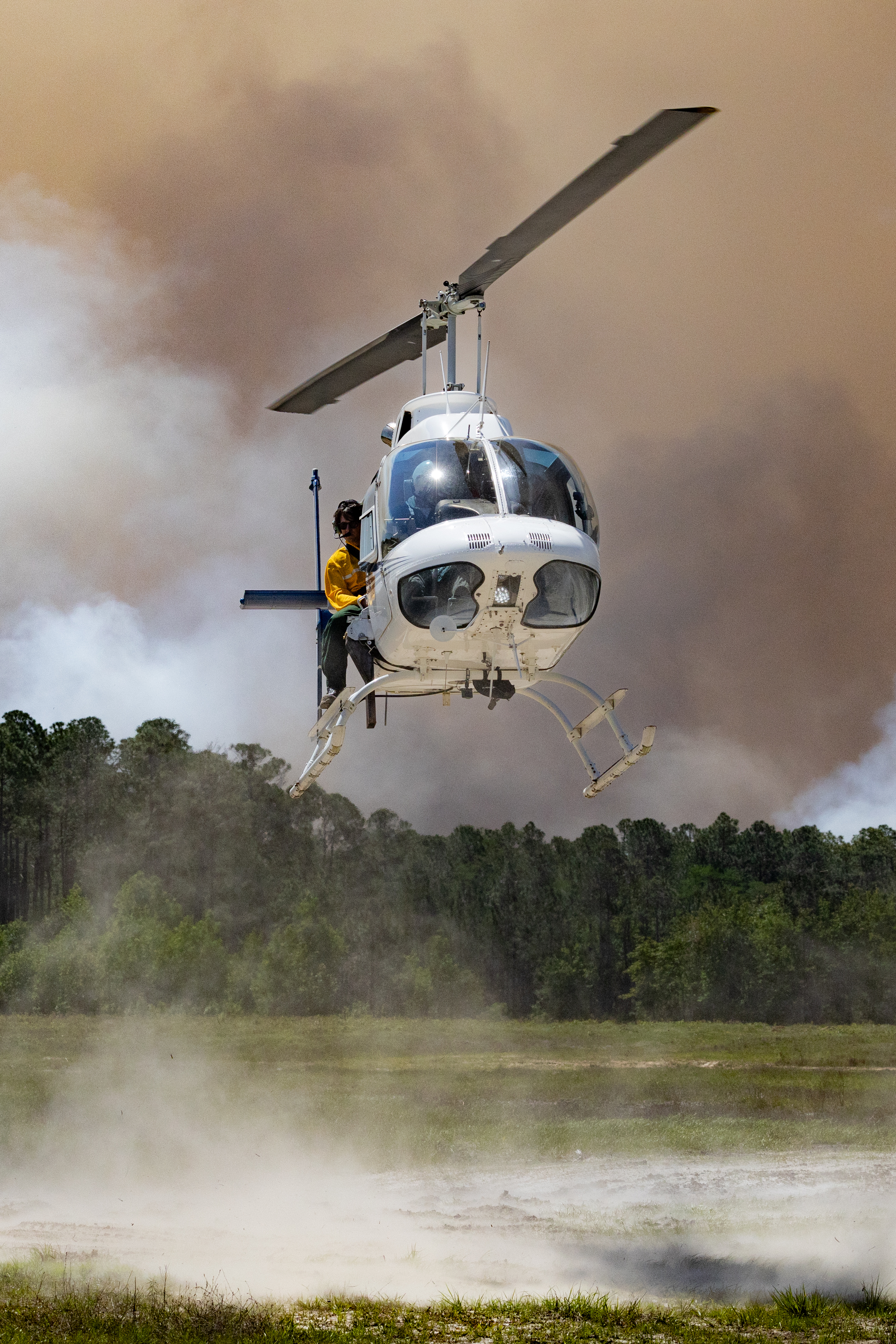 A white helicopter kicks up dust from a grassy clearing as it lands, against a backdrop of dark smoke rising over a green forest. A man in a yellow shirt leans out of the door.