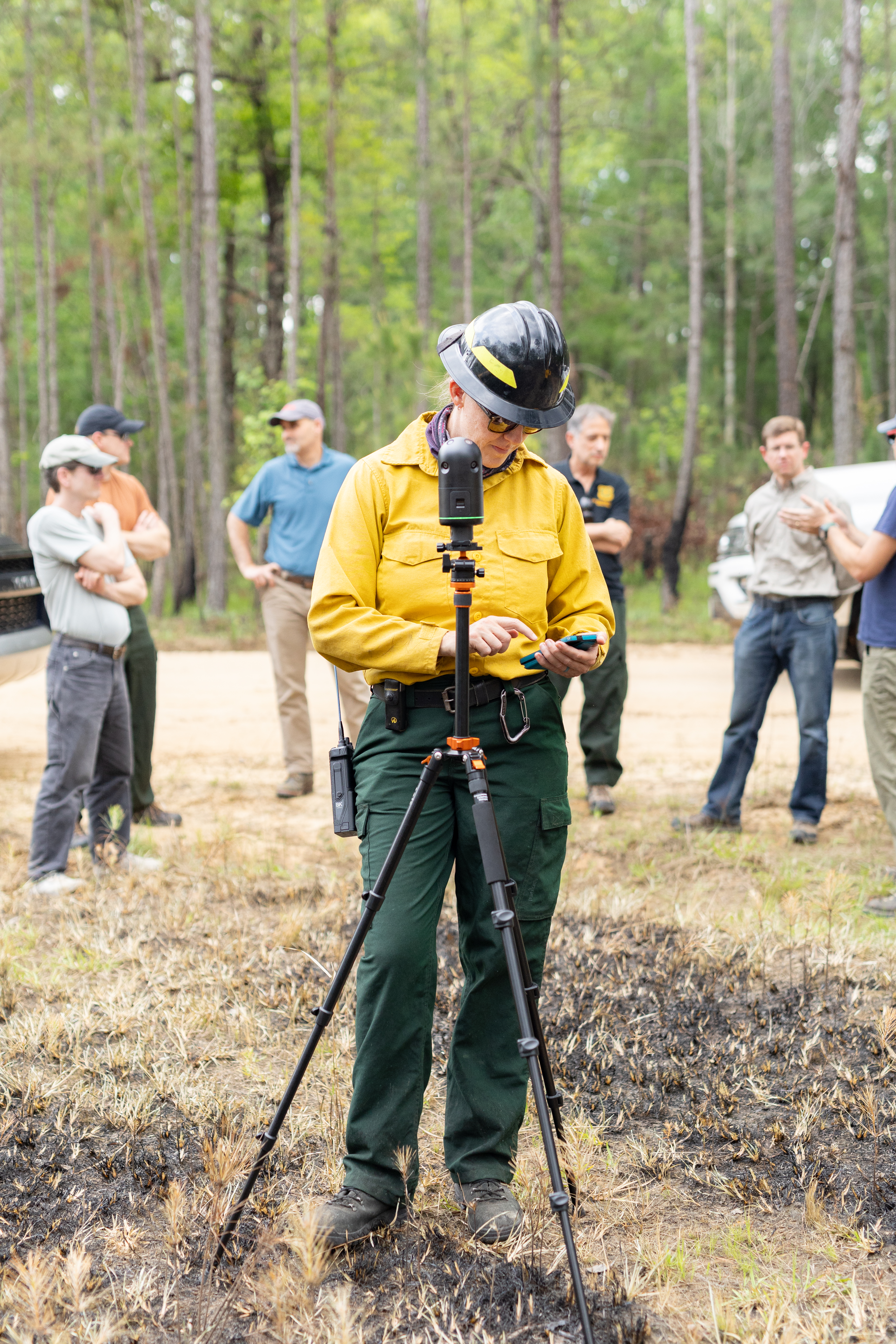 A woman in a yellow long-sleeved and black hard hat looks down at her phone, setting up a small device on a black tripod just in front of her. Behind her a group of about six people gather in a circle.