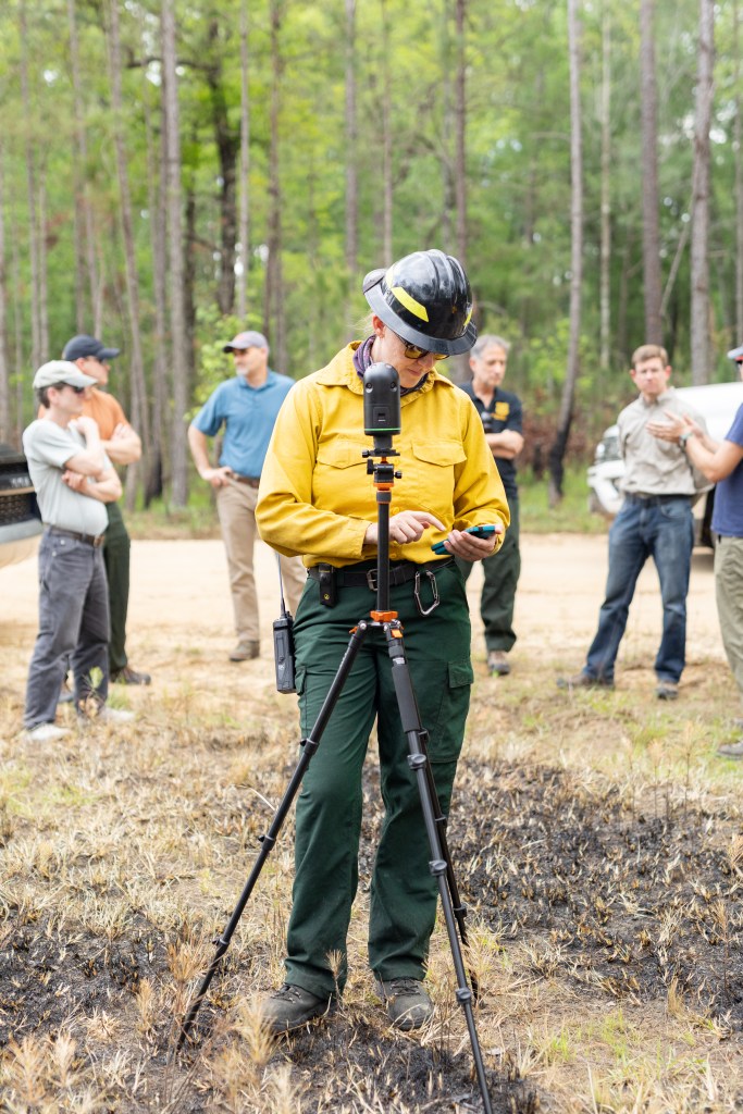A woman in a yellow long-sleeved and black hard hat looks down at her phone, setting up a small device on a black tripod just in front of her. Behind her a group of about six people gather in a circle.