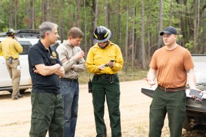 Three men speak with a woman in protective gear as she shows them something on her phone.