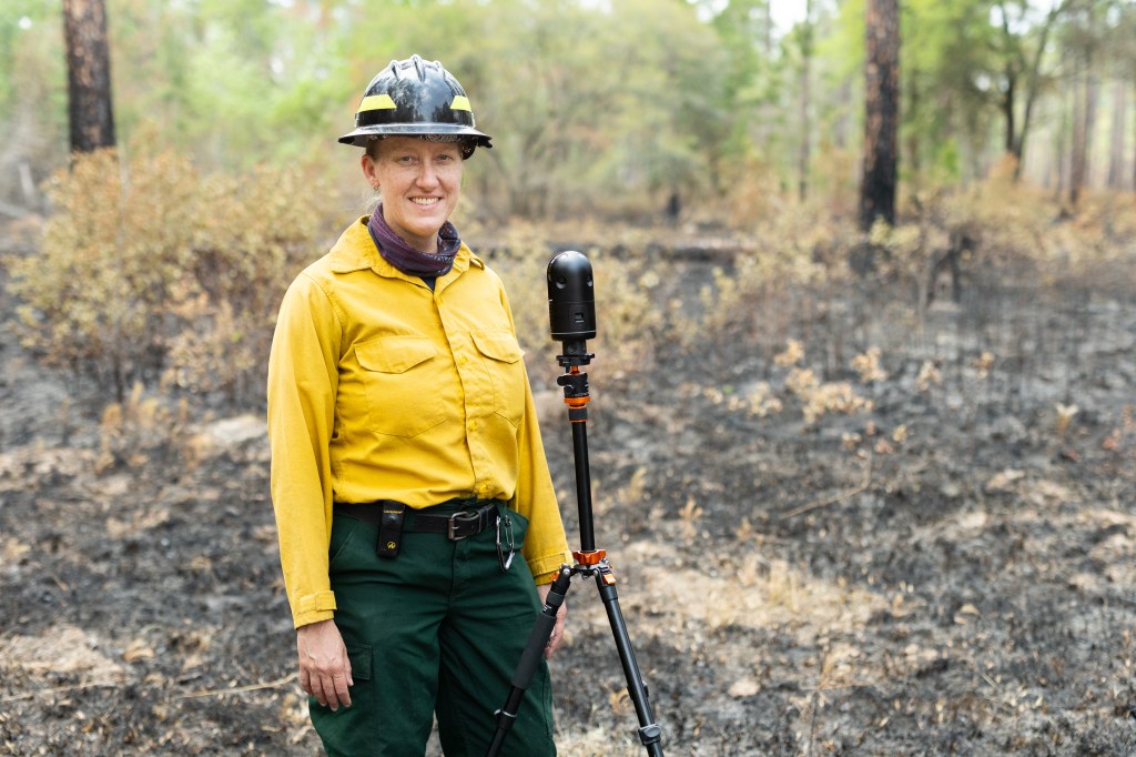 A woman in a yellow long-sleeved and black hard hat smiles next to a small device on a black tripod, in front of a forest.
