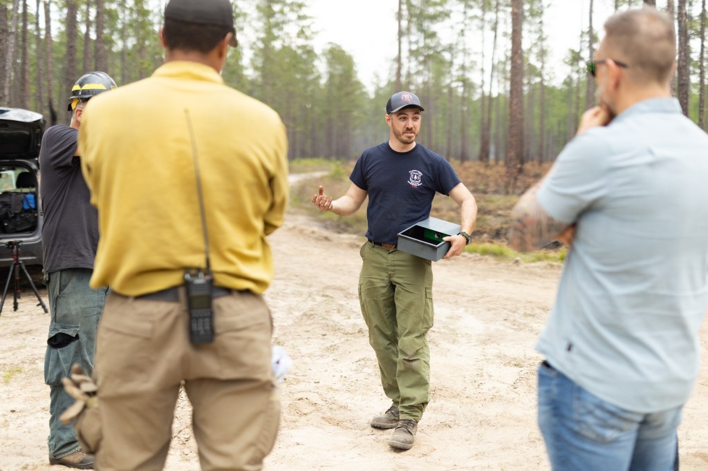 A man in a hat and navy t-shirt holds a metal box about the size of a lunchbox, gesturing to a few people with their backs to the camera.