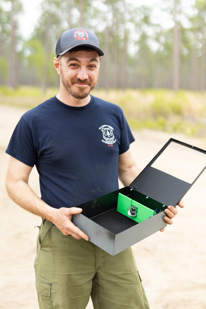 A man in a gray hat and navy t-shirt smiles while holding a metal box about the size of a lunchbox, with a small camera mounted on bright green plastic inside.