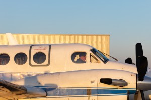 Golden sun illuminates the side of a white aircraft sitting on the tarmac, with the profile of a pilot visible through the window.