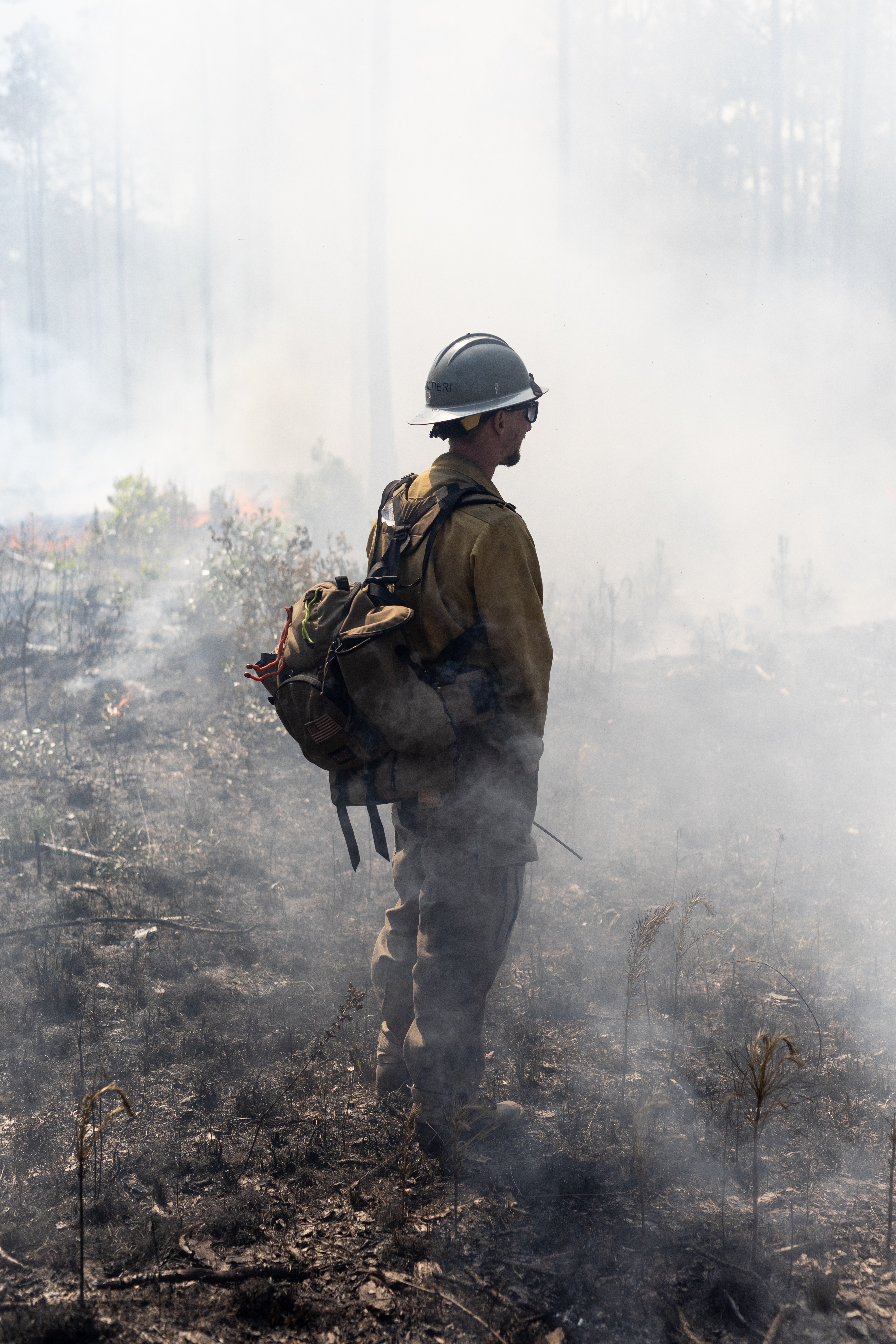 A man in dark clothing, wearing a white hard hat and brown backpack, stands on blackened ground, partially silhouetted against thick white smoke.