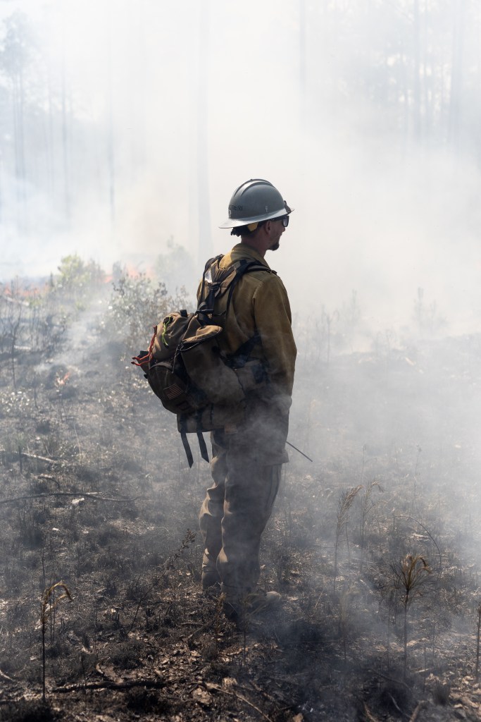 A man in dark clothing, wearing a white hard hat and brown backpack, stands on blackened ground, partially silhouetted against thick white smoke.