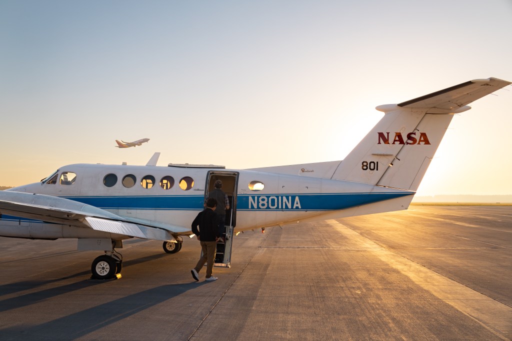 A man in black walks toward a white aircraft with a blue stripe, backlit by the gold of a rising sun. In the distance, an airplane takes off.