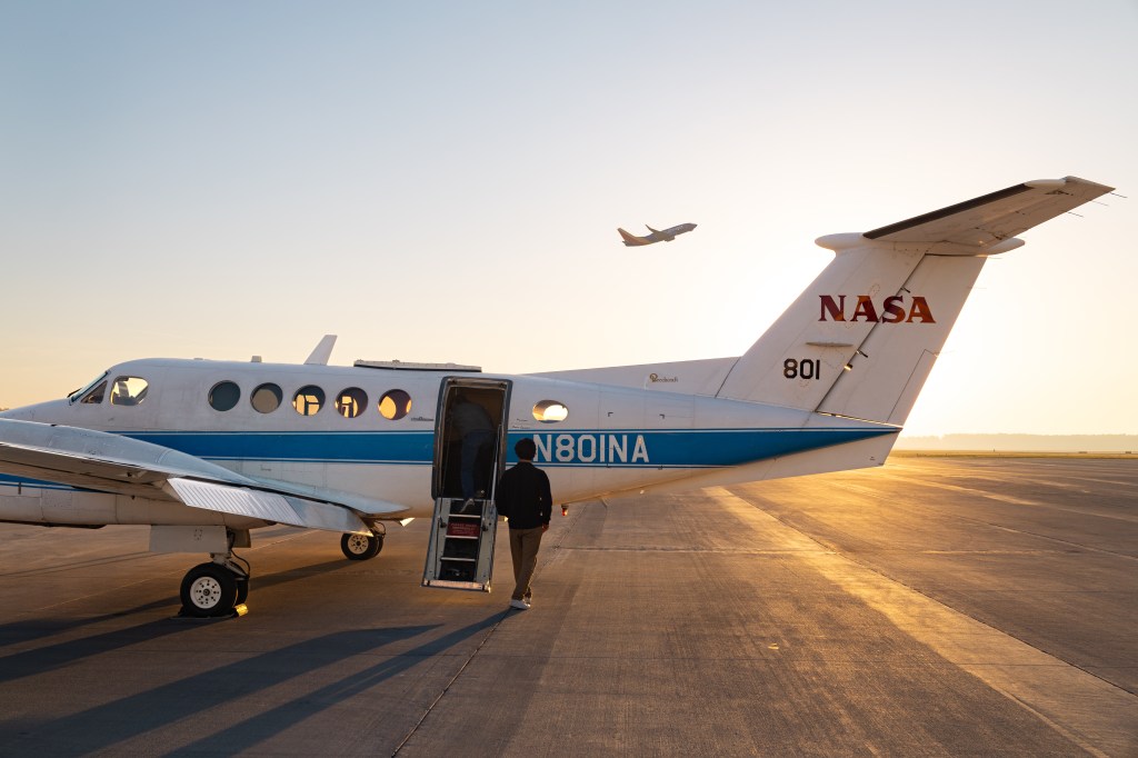 A man in black walks toward a white aircraft with a blue stripe, backlit by the gold of a rising sun. In the distance, an airplane takes off.