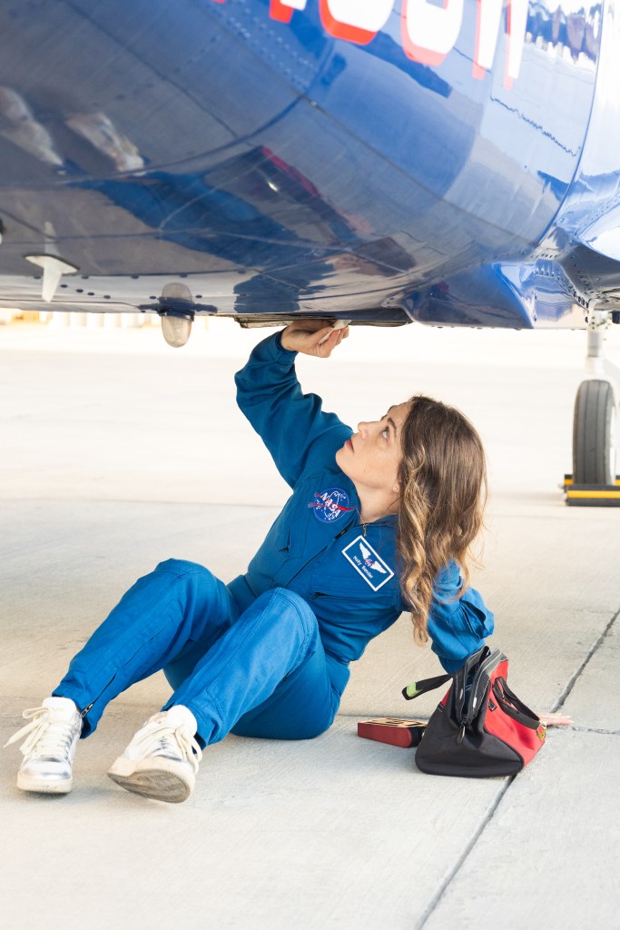 A woman in a blue NASA jumpsuit sits on gray tarmac beneath the blue underbelly of a plane, cleaning one of the ports.