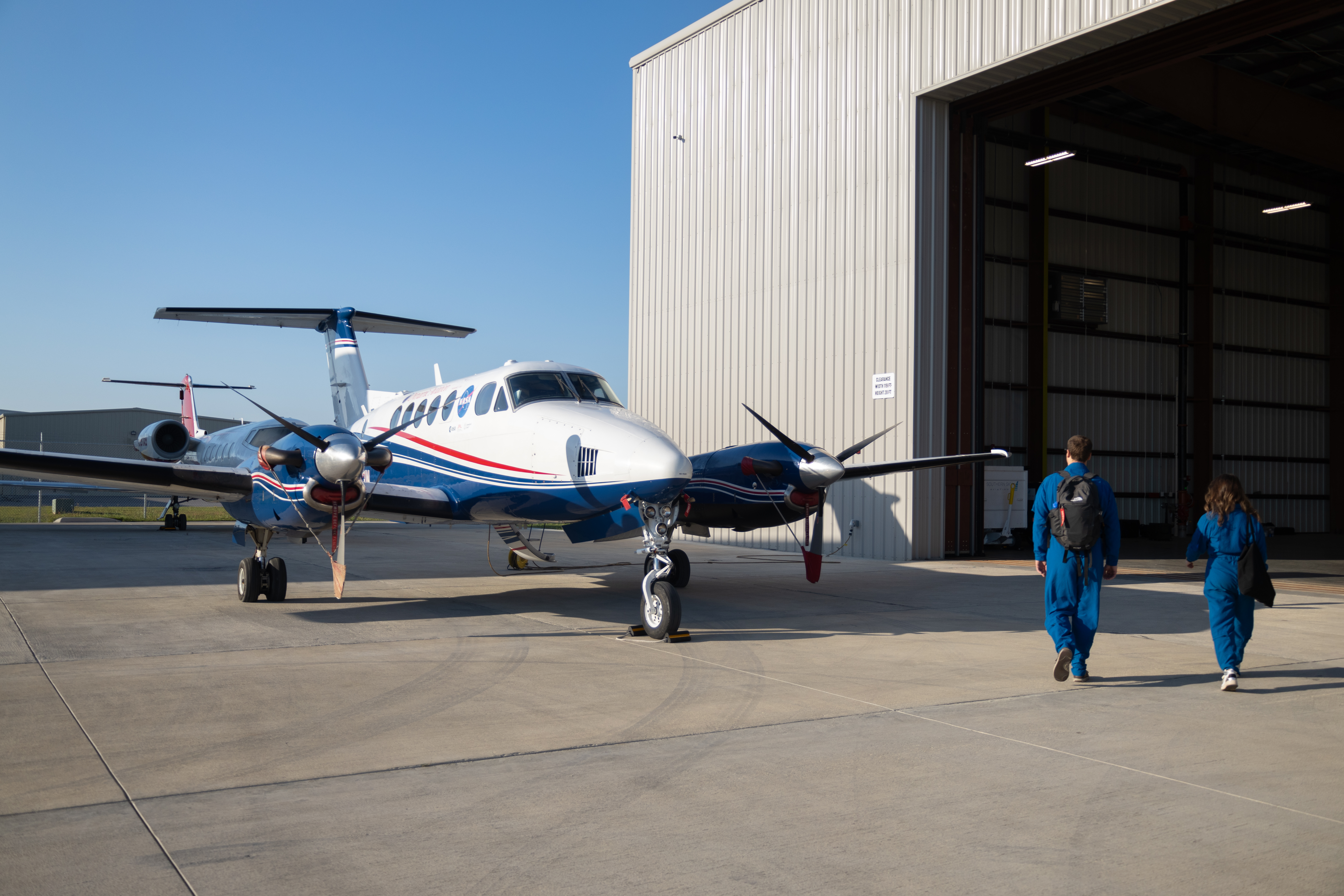 Two people in blue flight suits walk towards a small blue and white aircraft, sitting just outside a hangar on the tarmac.