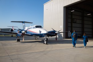 Two people in blue flight suits walk towards a small blue and white aircraft, sitting just outside a hangar on the tarmac.