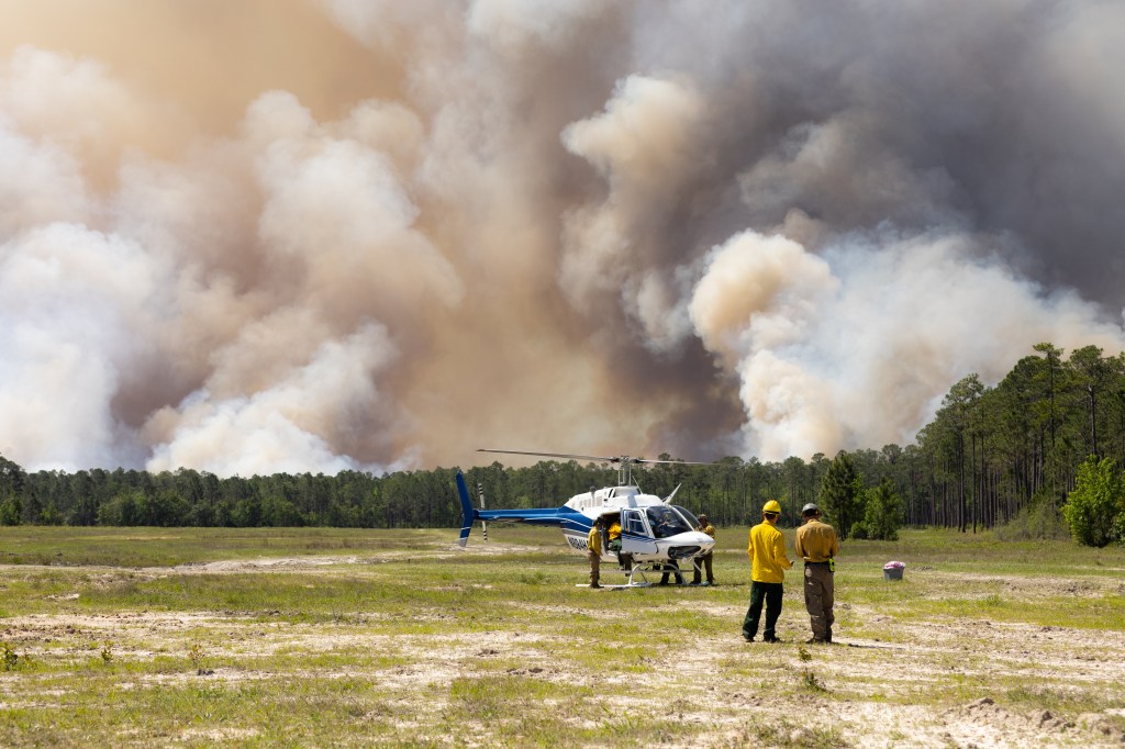 A white helicopter sits in a grassy clearing while dark gray and brown clouds of smoke rise above a treeline in the distance. In the foreground, two men in yellow shirts stand together looking down at something.