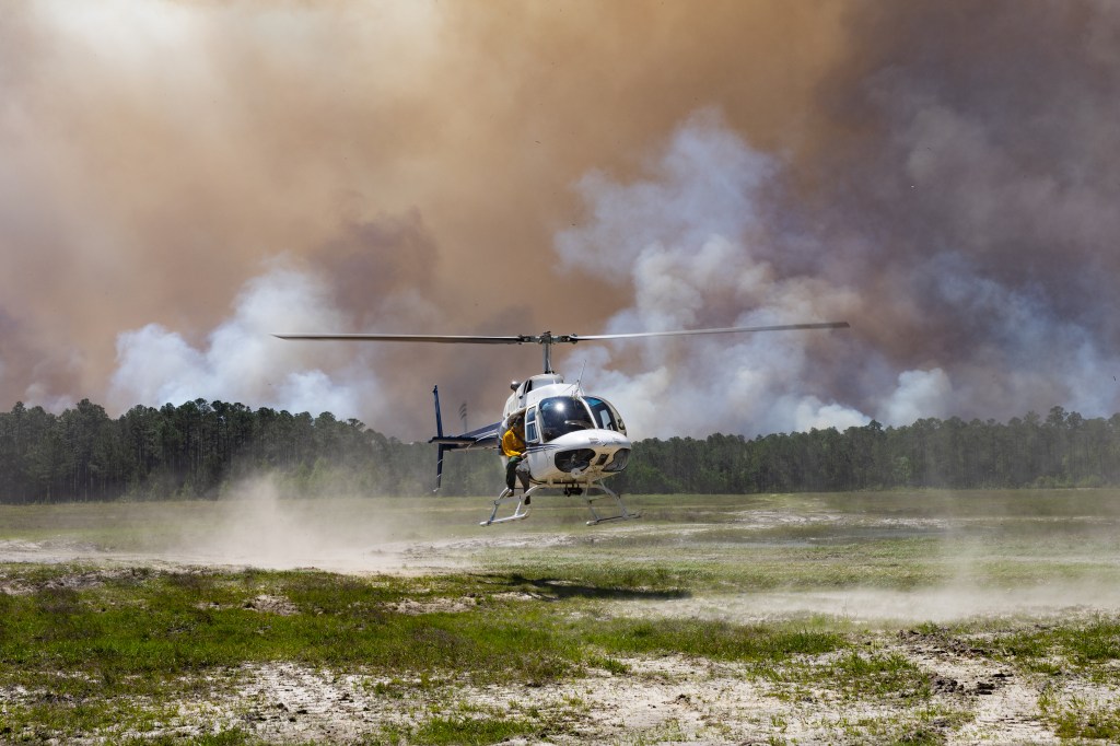 A white helicopter kicks up dust from a grassy clearing as it lands, against a backdrop of dark smoke rising over a green forest. A man in a yellow shirt leans out of the door.