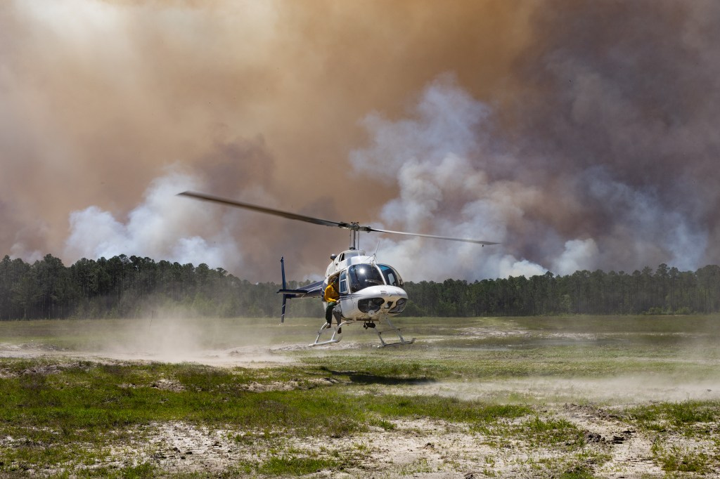 A white helicopter kicks up dust from a grassy clearing as it lands, against a backdrop of dark smoke rising over a green forest. A man in a yellow shirt leans out of the door.