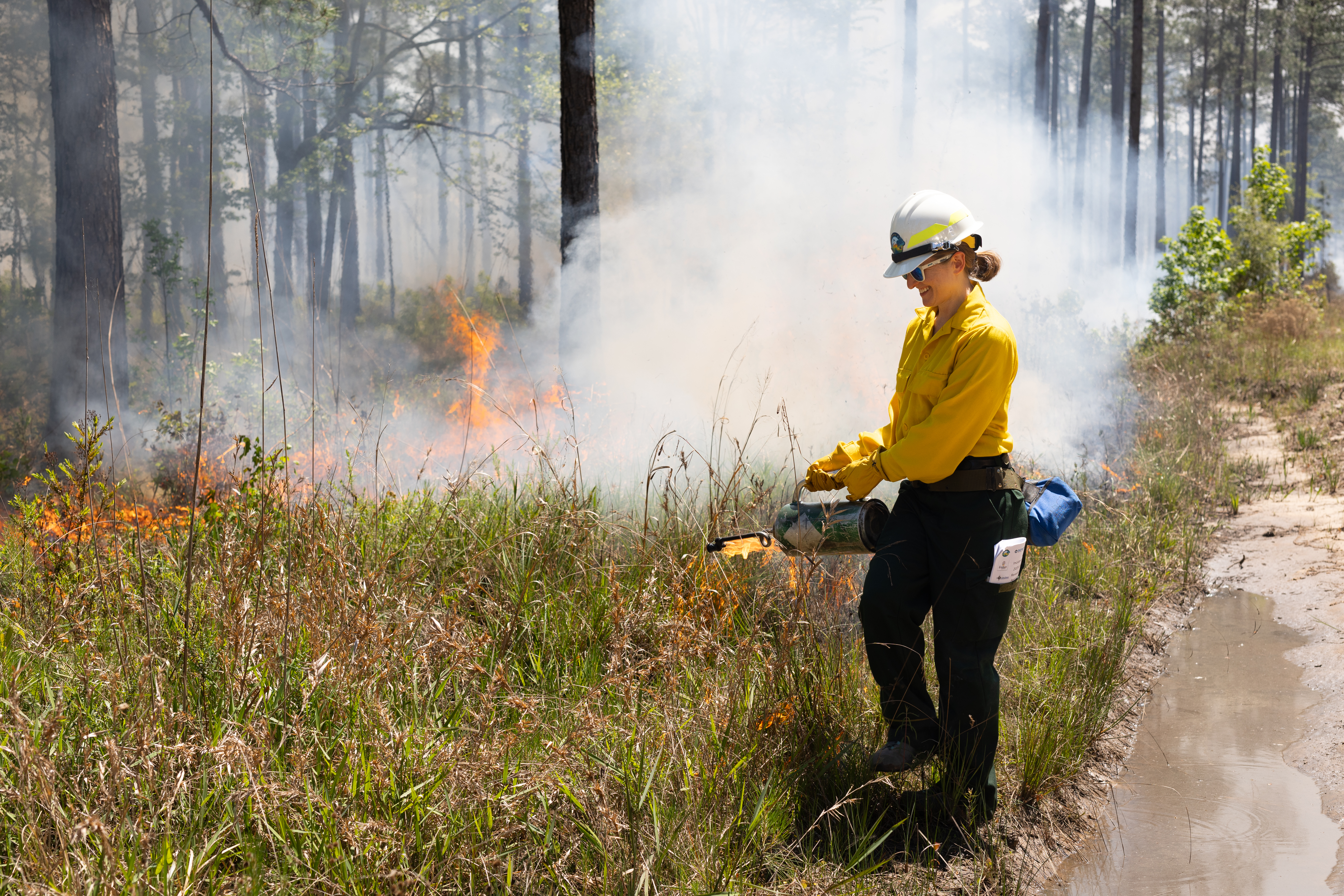 A woman in a yellow long-sleeved and protective gear holds a metal canister with a spout, dripping fire into green brush. In the background small flames send up a cloud of white smoke.
