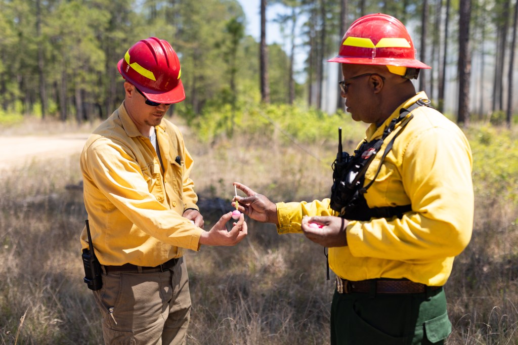 Two firefighters in yellow long-sleeved shirts and red hardhats perform a demo: the one on the left holds a small plastic sphere, while the one on the right injects it with a small syringe.