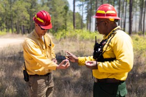 Two firefighters in yellow long-sleeved shirts and red hardhats perform a demo: the one on the left holds a small plastic sphere, while the one on the right injects it with a small syringe.