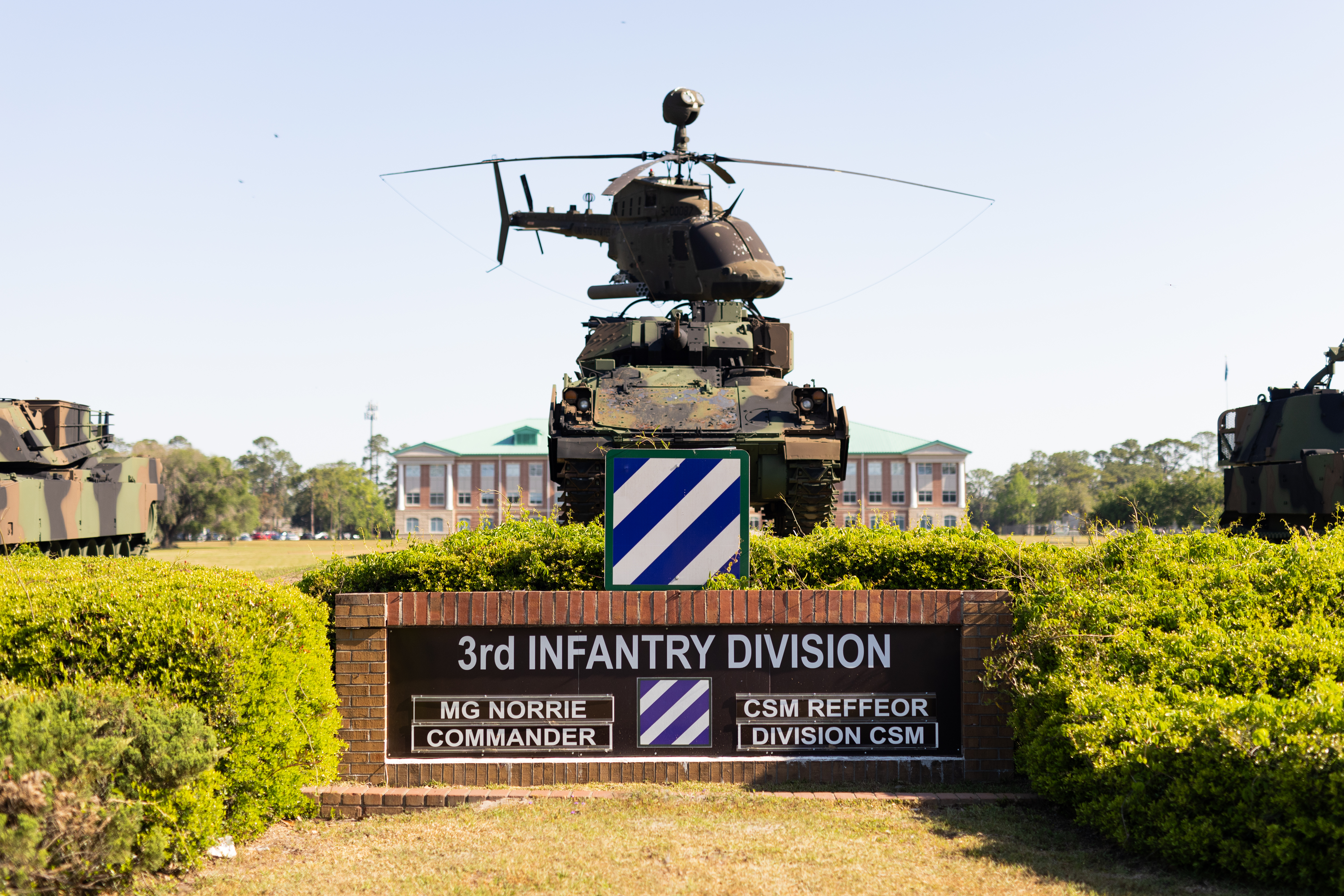 A sign that reads 3rd Infantry Division is rimmed in red brick, surrounded by green hedges. Above the sign, models of a tank and a helicopter sit poised and and painted in camo.