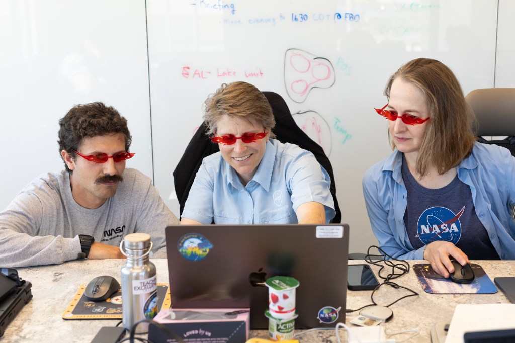 Three people sit at a table, all looking at a laptop with a whiteboard behind them. From left: a man with short dark hair and a large mustache, a woman with short sandy hair, and a woman with medium-length sandy hair. All three wear red sunglasses shaped like flames.