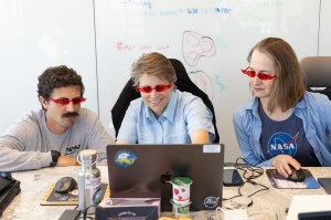 Three people sit at a table, all looking at a laptop with a whiteboard behind them. From left: a man with short dark hair and a large mustache, a woman with short sandy hair, and a woman with medium-length sandy hair. All three wear red sunglasses shaped like flames.