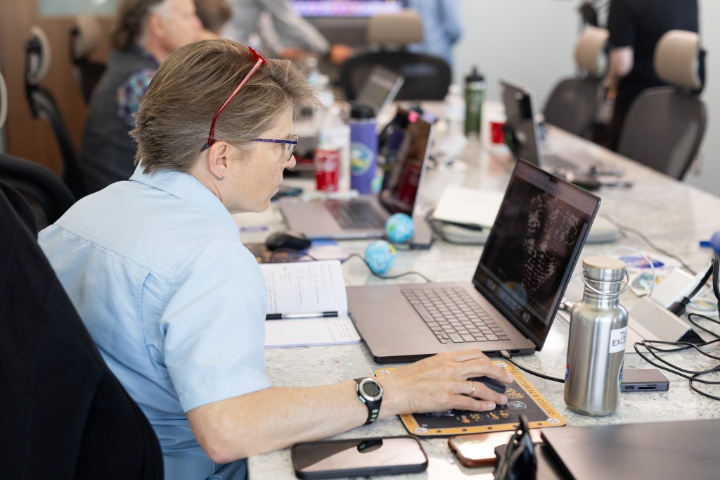 A woman in a blue shirt with red sunglasses on her head sits at a table, looking at a laptop.