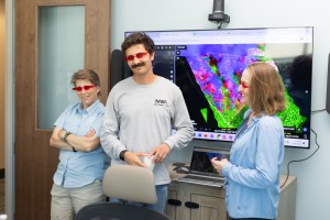 Three people stand in front of a monitor displaying data points in neon green, pink, and purple. From left: a woman with short sandy hair, a man with short dark hair and a large mustache,and a woman with medium-length sandy hair. All three wear red sunglasses shaped like flames.