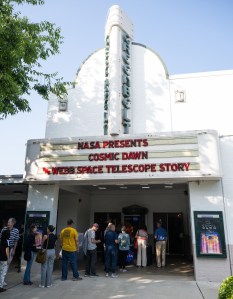 10 people stand in a line at a movie theater. About half of the people are standing under the movie marquee, which reads "NASA presents Cosmic Dawn The Webb Space Telescope Story" in red capital letters.