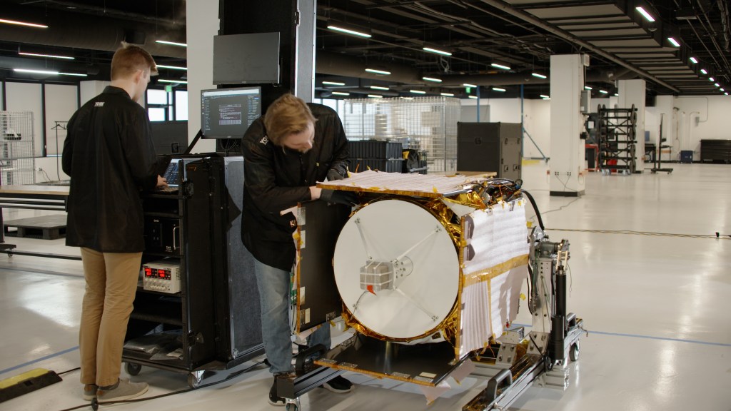 In a large white room within Johns Hopkins Applied Physics Laboratory, two engineers in black York Space Systems coats monitor the Polylingual Experimental Terminal (PExT). The terminal is mounted horizontally on a wheeled metal cart, its white antenna dish pointed outward. The payload is enclosed in white insulation and gold tape. One technician leans over the terminal to investigate PExT’s antenna while the other stands at a laptop to the left of it, reviewing data on a testing monitor.
