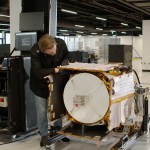 In a large white room within Johns Hopkins Applied Physics Laboratory, two engineers in black York Space Systems coats monitor the Polylingual Experimental Terminal (PExT). The terminal is mounted horizontally on a wheeled metal cart, its white antenna dish pointed outward. The payload is enclosed in white insulation and gold tape. One technician leans over the terminal to investigate PExT’s antenna while the other stands at a laptop to the left of it, reviewing data on a testing monitor.