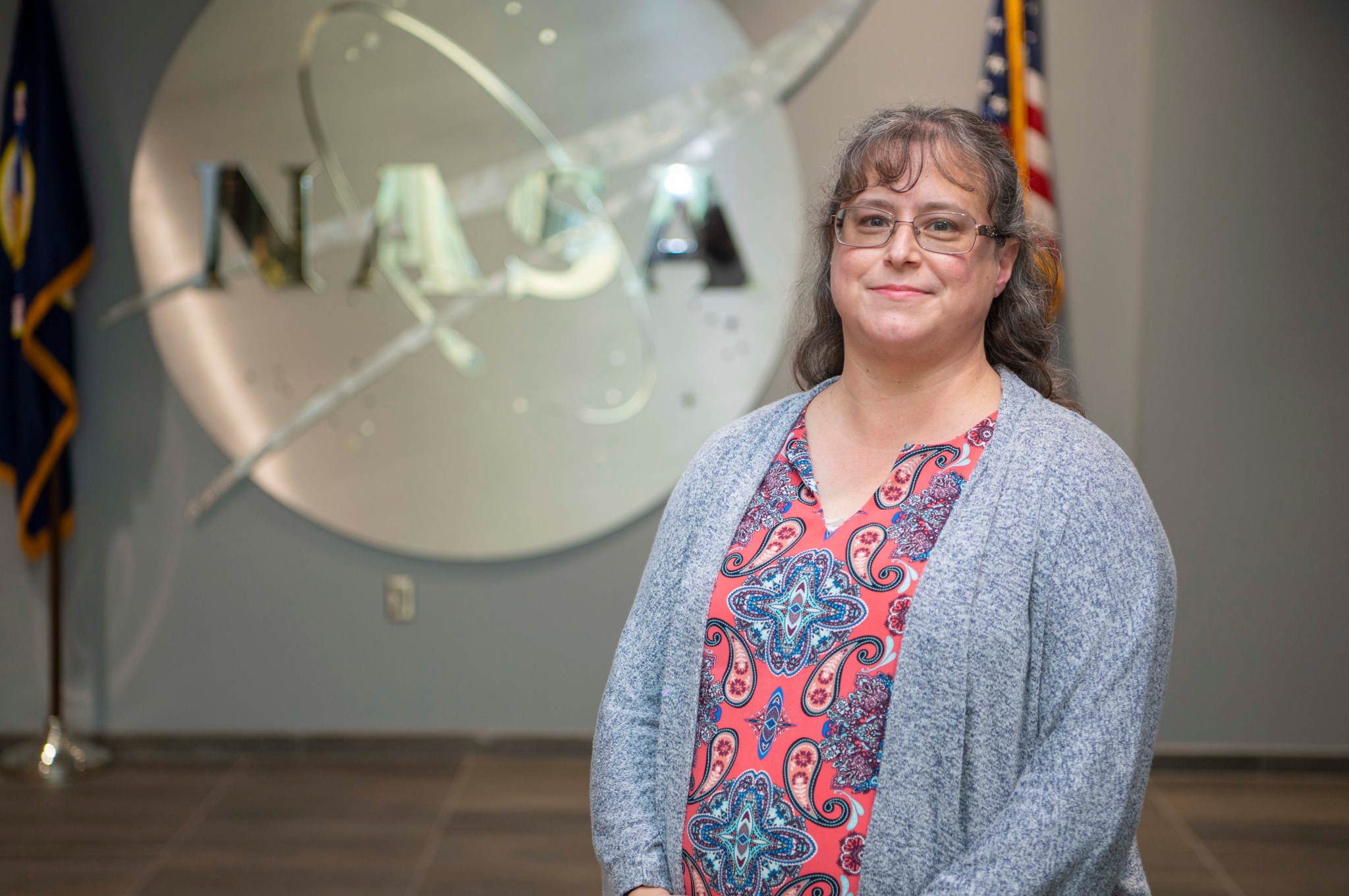Christina Zeringue wearing a gray cardigan stands before a silver NASA insignia