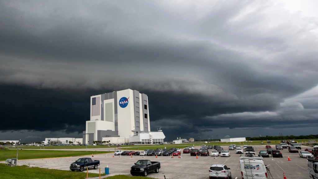 A large front of dark gray storm clouds looms over a light gray cement building with the NASA meatball on the side. In the foreground is a parking lot.