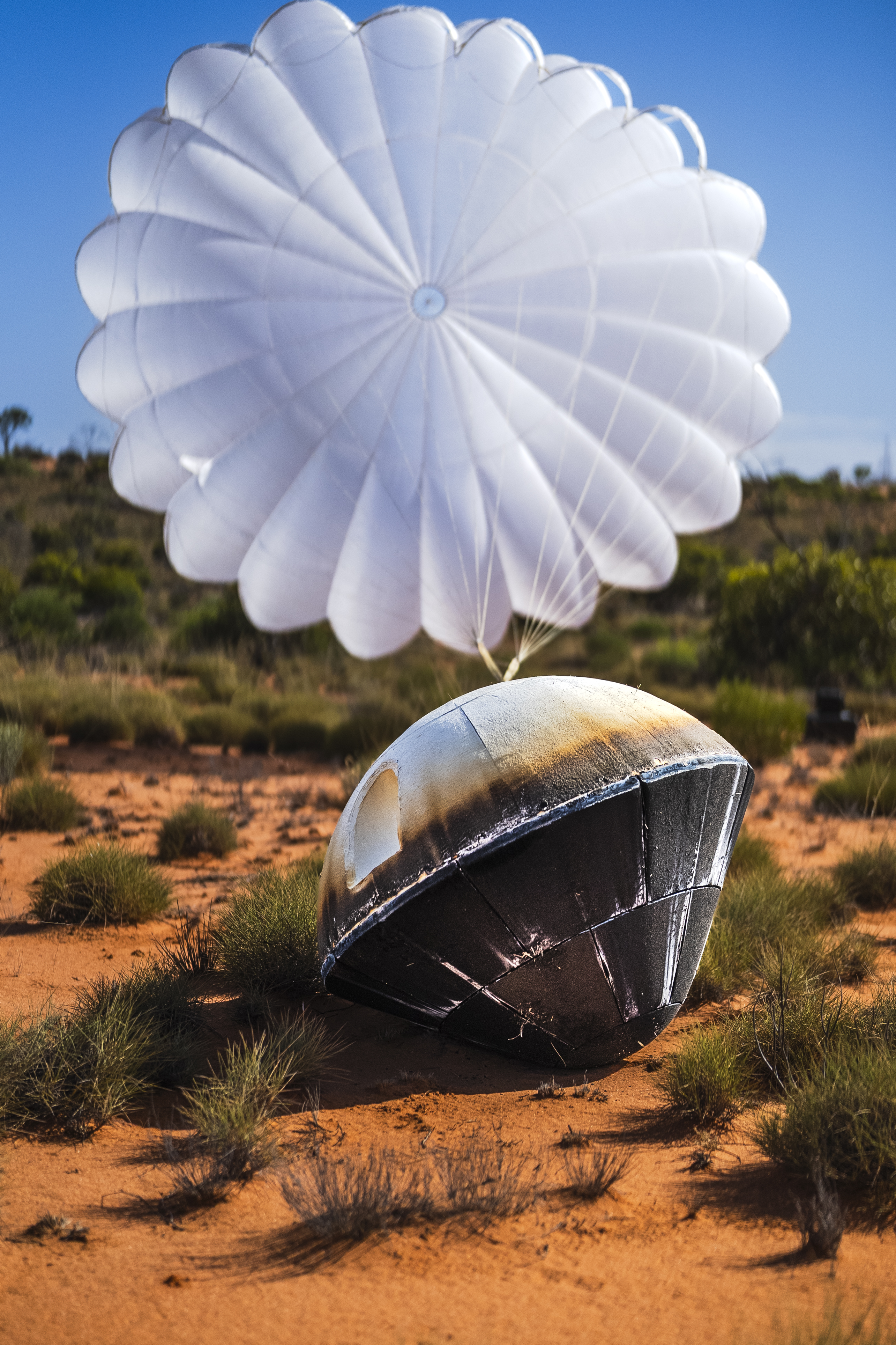 A capsule with burn marks and a billowing parachute behind it sits on the desert floor.