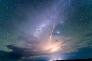 A light streaks across a starry night sky lit at the horizon and illuminating wispy clouds.
