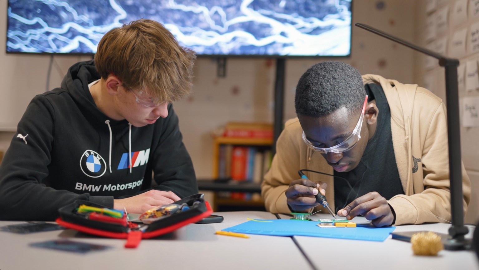 Two high school students sit at a work bench soldering.