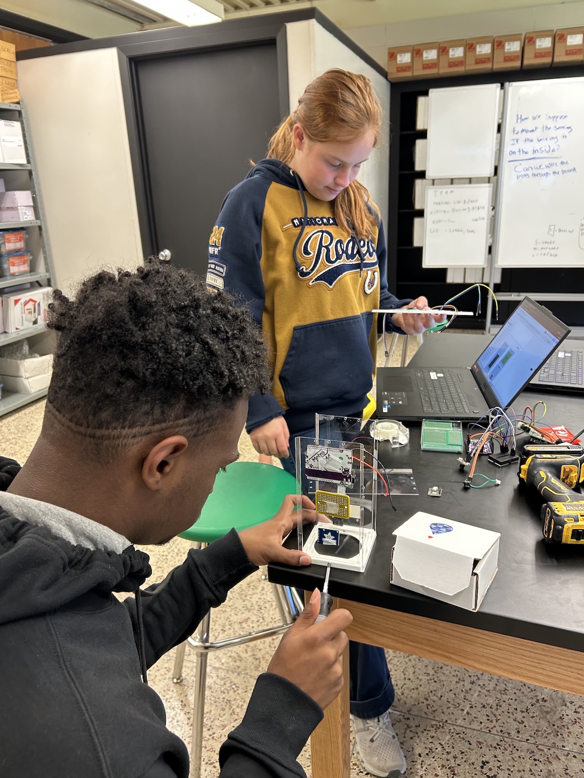 A student sits at a work table unscrewing a fastner holding an experiment in its case while another student looks on.
