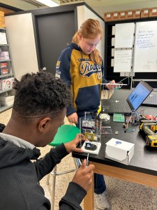 A student sits at a work table unscrewing a fastner holding an experiment in its case while another student looks on.