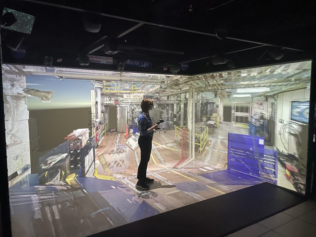 A girl wearing a VR headset and holding a controller in her hand gazes at a 3D environment of a wind tunnel facility. The walls around her and the floor beneath her are screens and display parts of this visualization.
