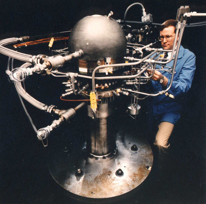 A technician inspects the 12.5-kW Component Test Power Convertor with a spherical core and attached fluid and electrical lines.