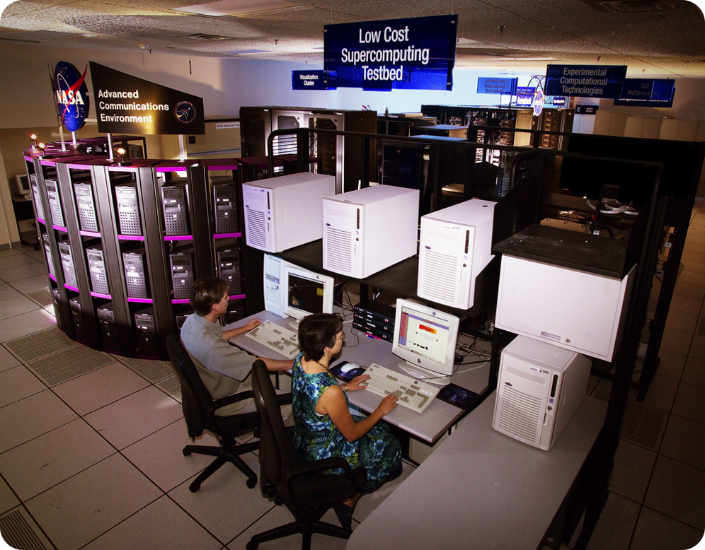 A man and a woman sit on rolling chairs at a table, typing on the large, blocky computers in front of them. Behind the table is a large section of computer stacked on top of one another, along with other various technology and equipment.
