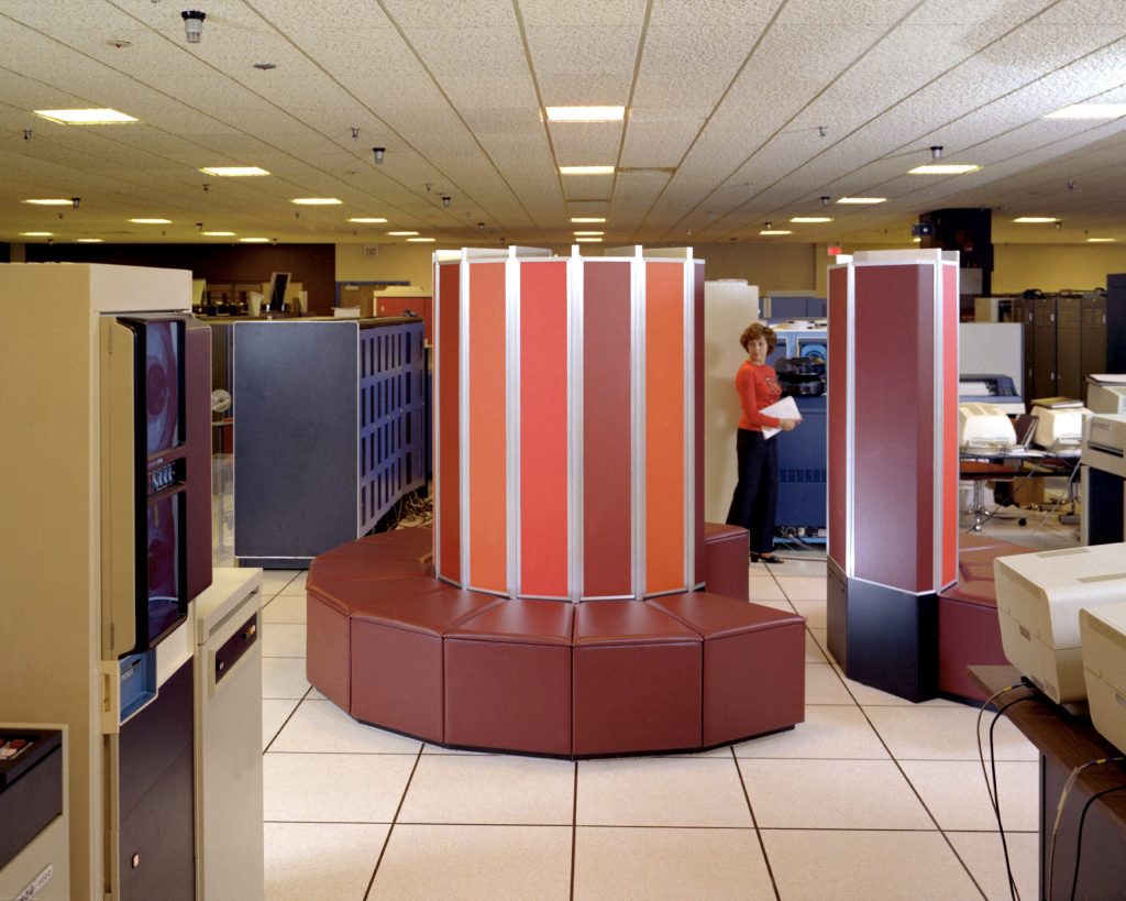 A large warm-colored rectangular device sits in a beige office room, surrounded by other cool-colored equipment and furniture.  A woman stands behind the large device.