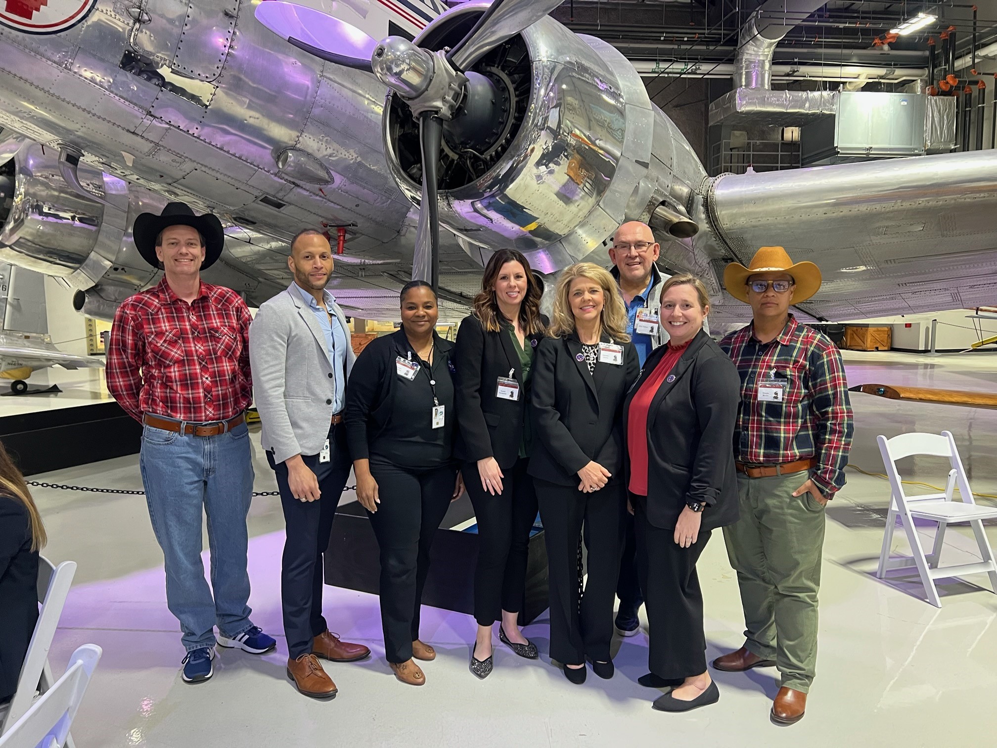 A group of eight men and women in business casual attire stand in front of a shiny silver aircraft.