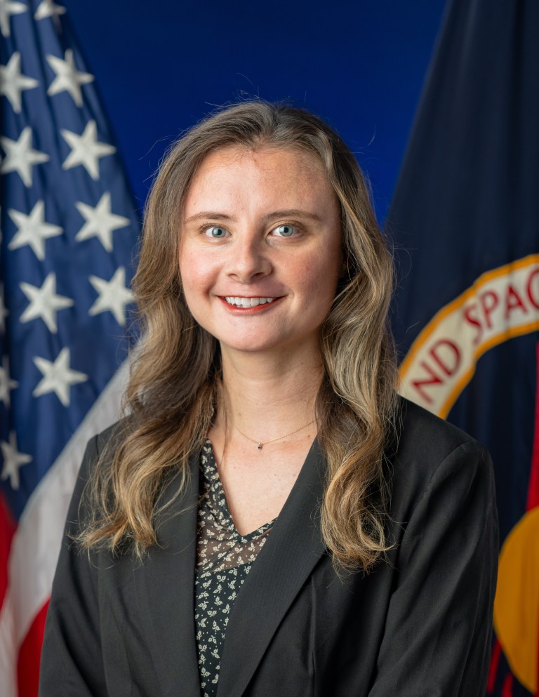 Headshot of Devan Nisson wearing a black blazer sitting in front of an American flag background