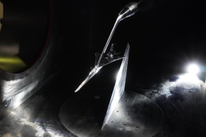 A scaled model of the X-59 experimental aircraft is inside a wind tunnel for testing. All of the visible surfaces and the plane are a metallic silver. The aircraft has a distinctive long nose with a curved end. A spotlight on the floor in the far corner lights up the scene.